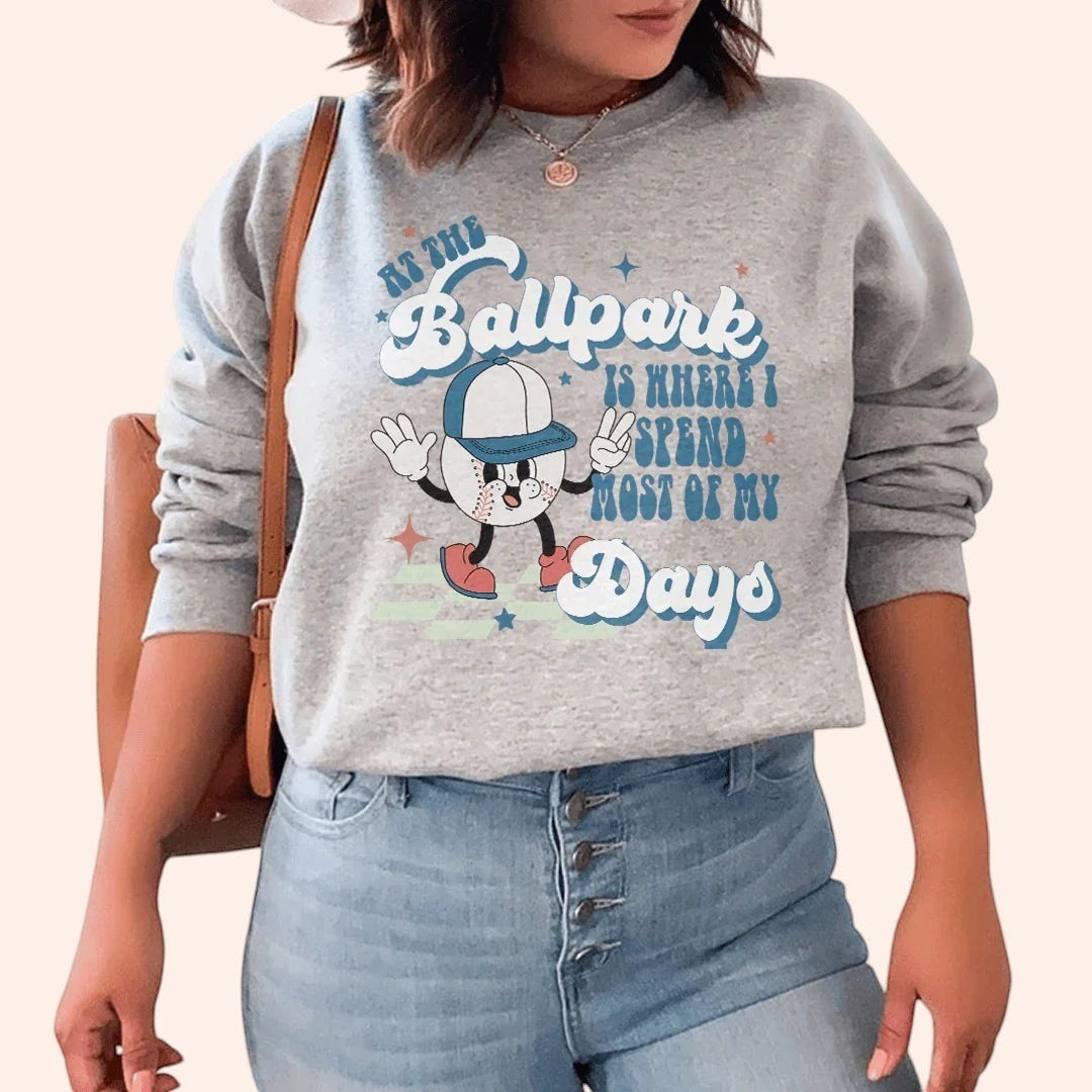Bleacher Babe grey crewneck sweatshirt with a cartoon baseball graphic and text reading "At the Ballpark Is Where I Spend Most of My Days."