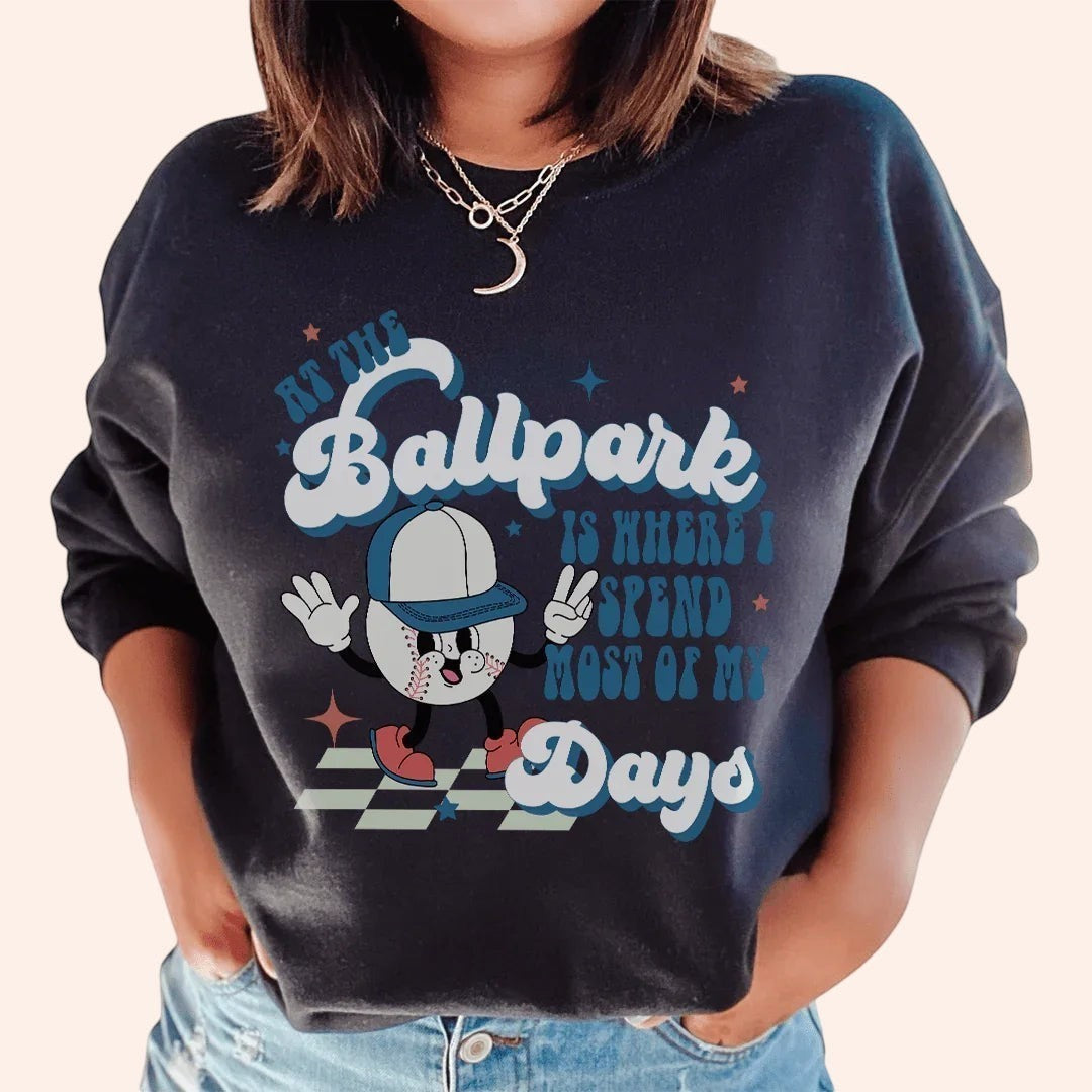 Bleacher Babe dark crewneck sweatshirt with a retro graphic of a baseball character and text "At the Ballpark Is Where I Spend Most of My Days."