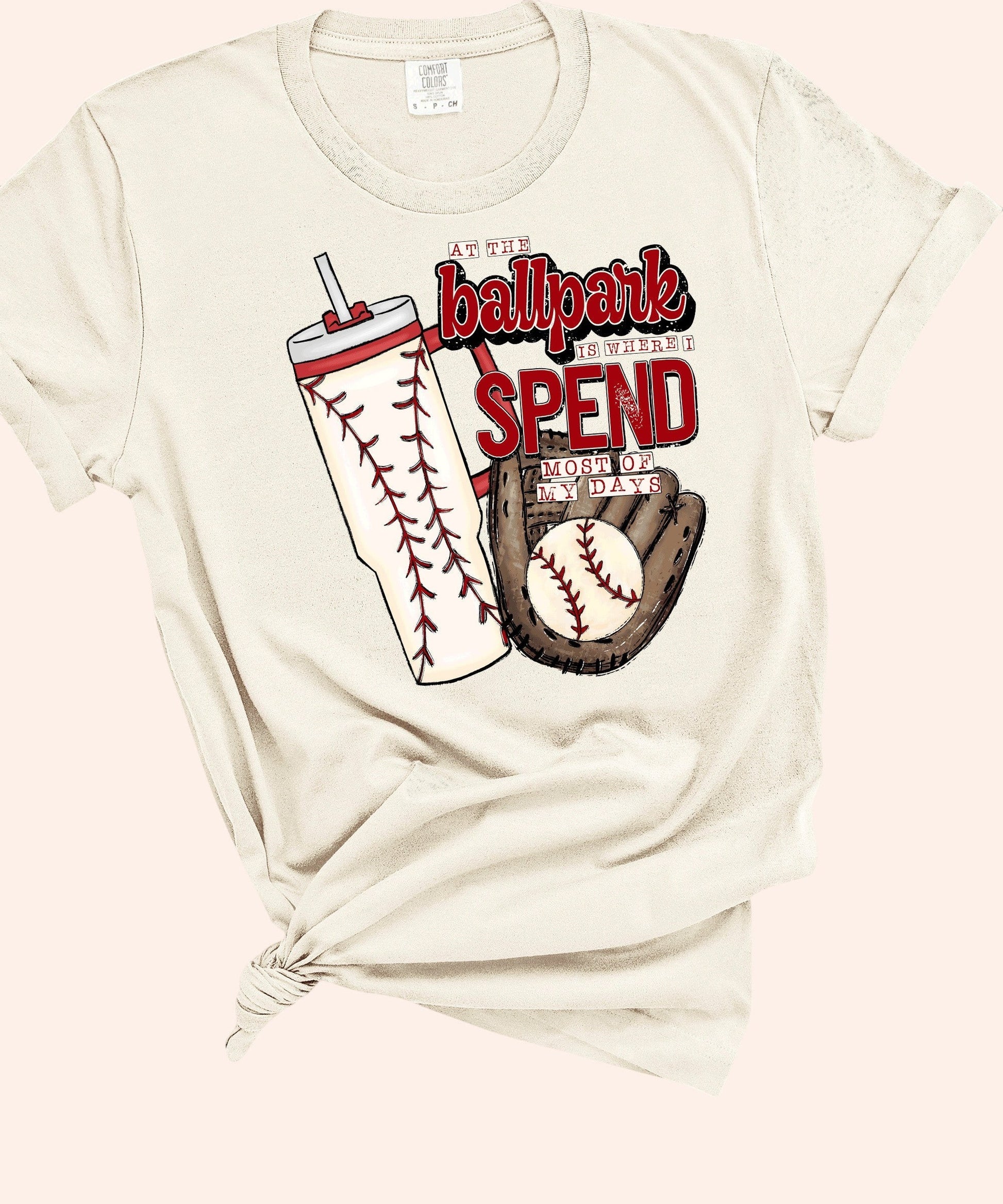Bleacher Babe baseball mom graphic tee in cream, featuring a tumbler with baseball stitching, a baseball glove, and the text "At the ballpark is where I spend most of my days."