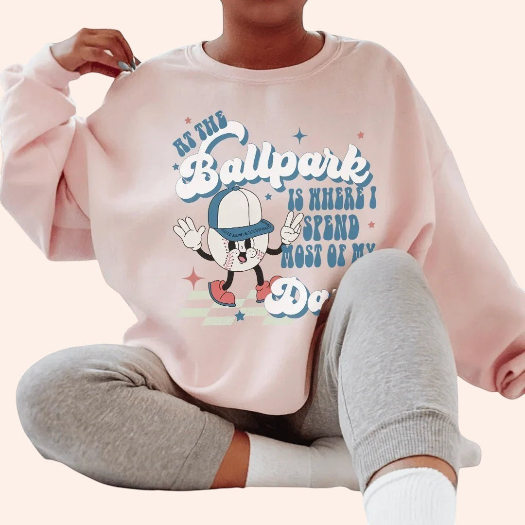 Bleacher Babe pink crewneck sweatshirt with a graphic of a cartoon baseball player and text "At the Ballpark Is Where I Spend Most of My Days."