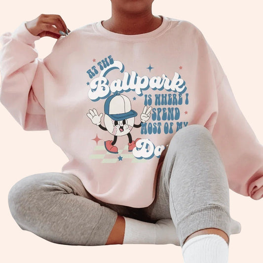 Bleacher Babe pink crewneck sweatshirt with a graphic of a cartoon baseball player and text "At the Ballpark Is Where I Spend Most of My Days."