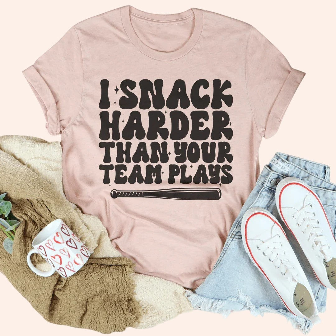 Bleacher Babe pink graphic tee with black text that reads "I Snack Harder Than Your Team Plays" and a baseball bat graphic.