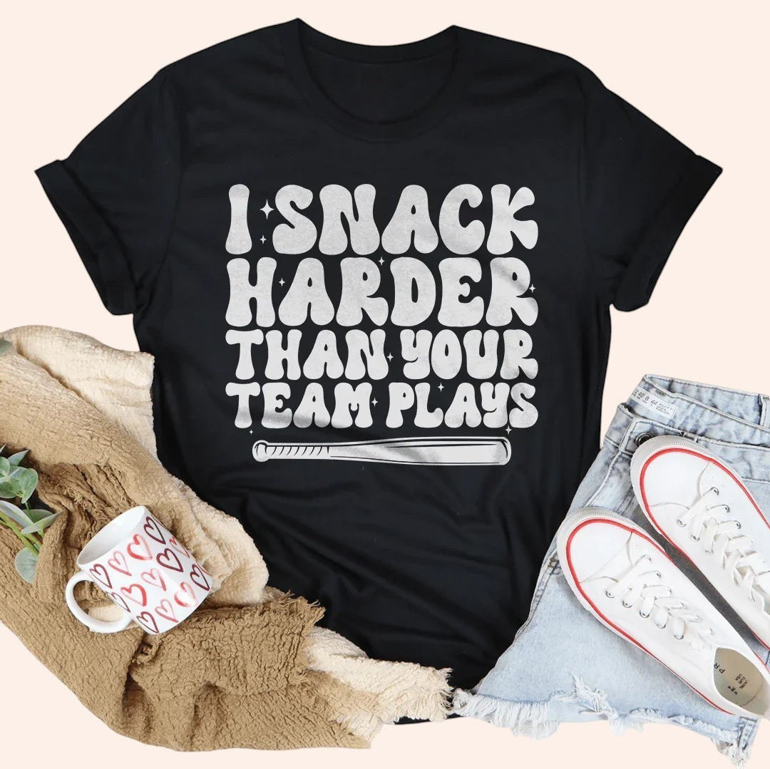 Bleacher Babe black cotton t-shirt with white text that reads "I Snack Harder Than Your Team Plays" and a baseball bat graphic.